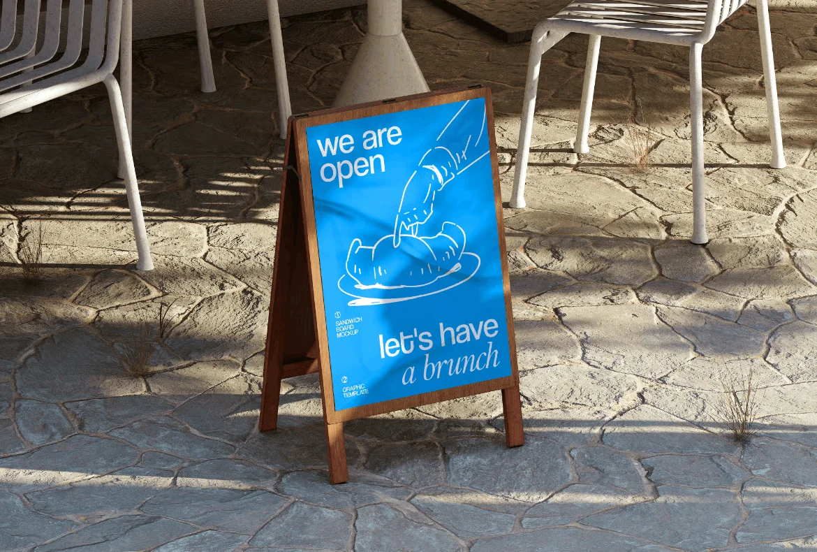 A blue Simpl by Rob&Paul sandwich board sign on a stone patio reads, We are open, let's have a brunch, with a white line drawing of a hand cutting into a croissant. White chairs and a table sit in the background.