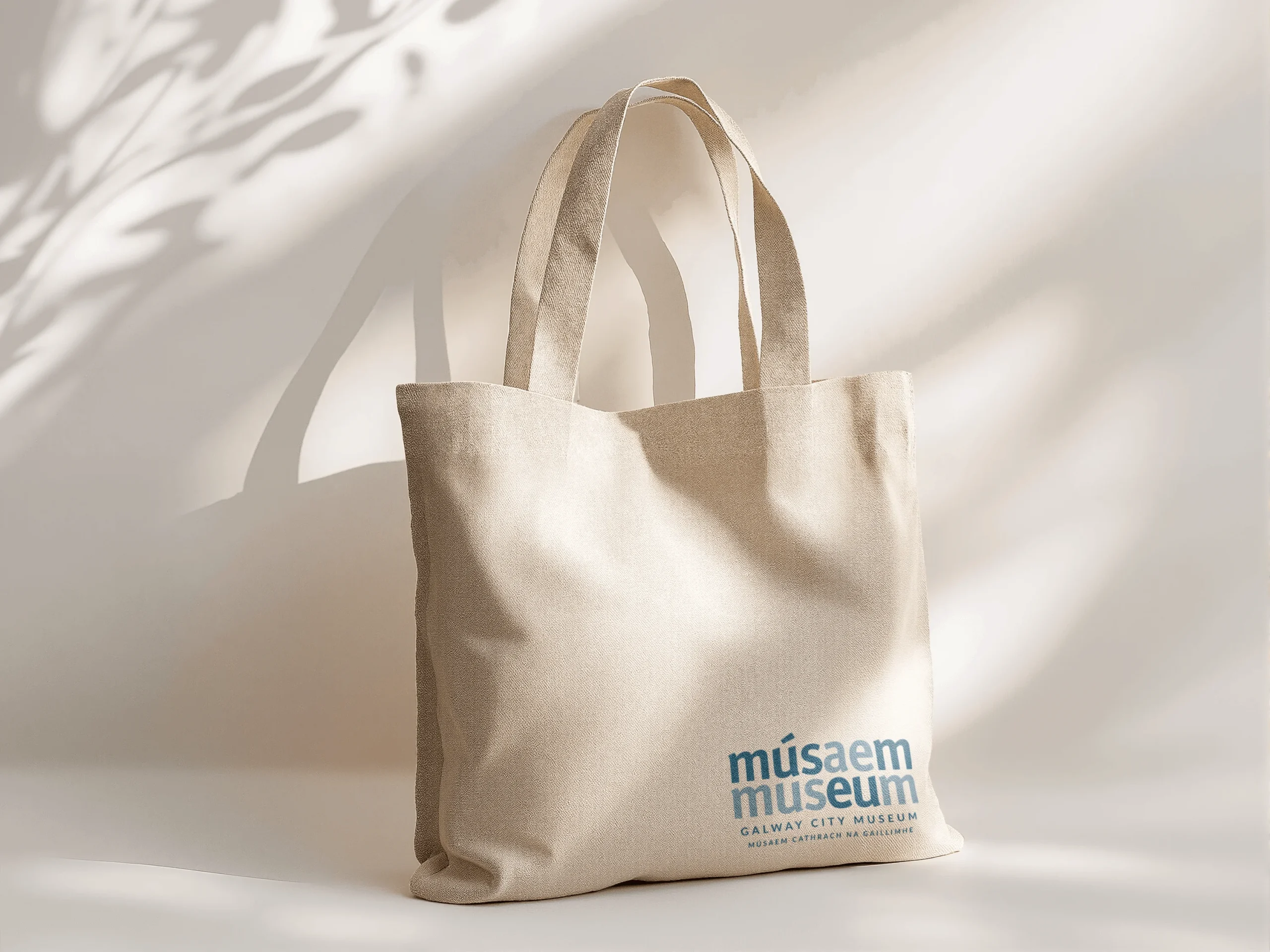 A beige fabric tote bag by Simpl by Rob&Paul with “músaem museum Galway City Museum” printed in blue on the lower right, standing upright as soft natural light casts leaf shadows on the background.