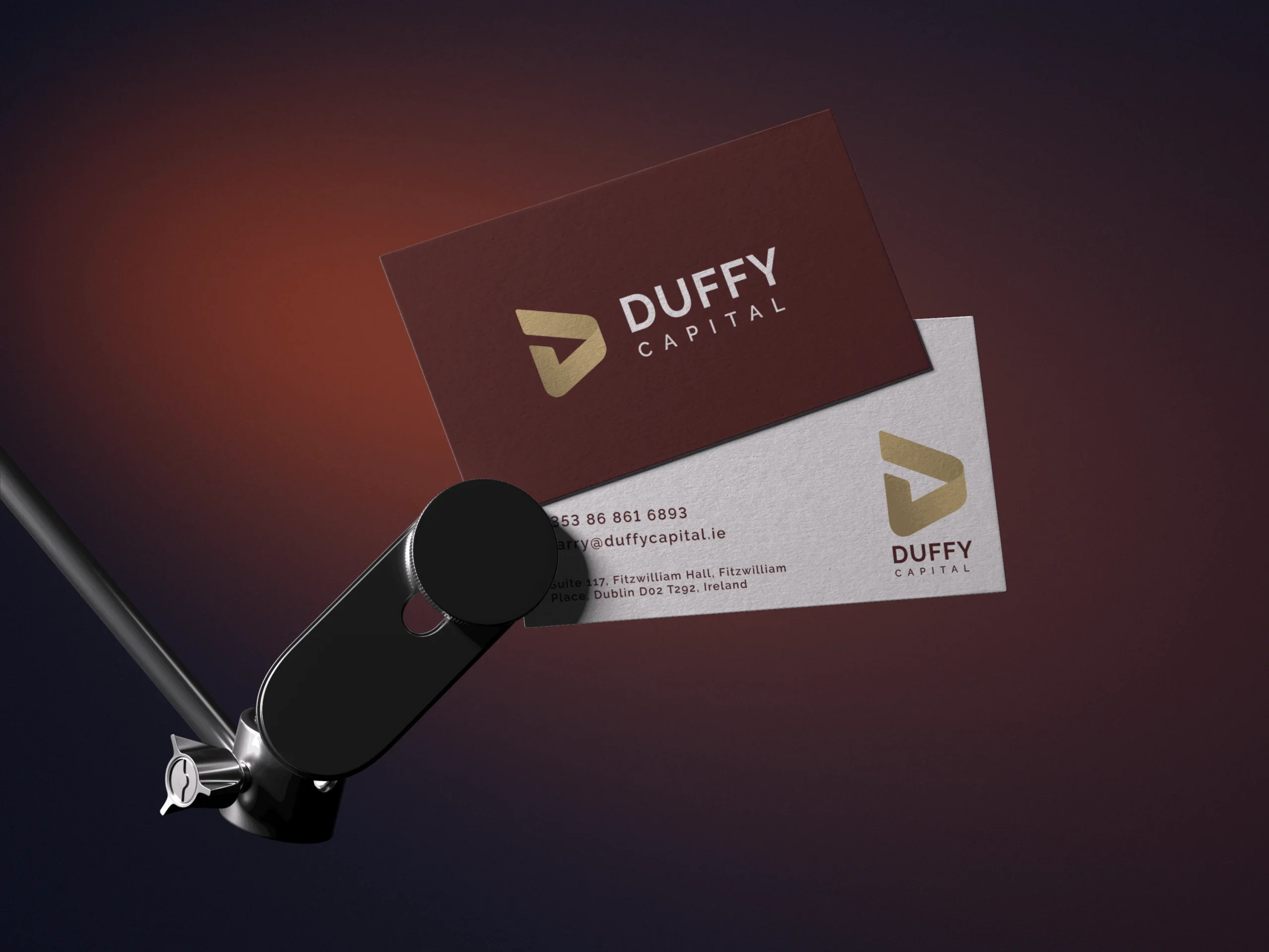 Two business cards for Duffy Capital, designed with Simpl by Rob&Paul, are held by a black clamp against a dark gradient background—one maroon with gold logo and text, the other white with black and gold details.