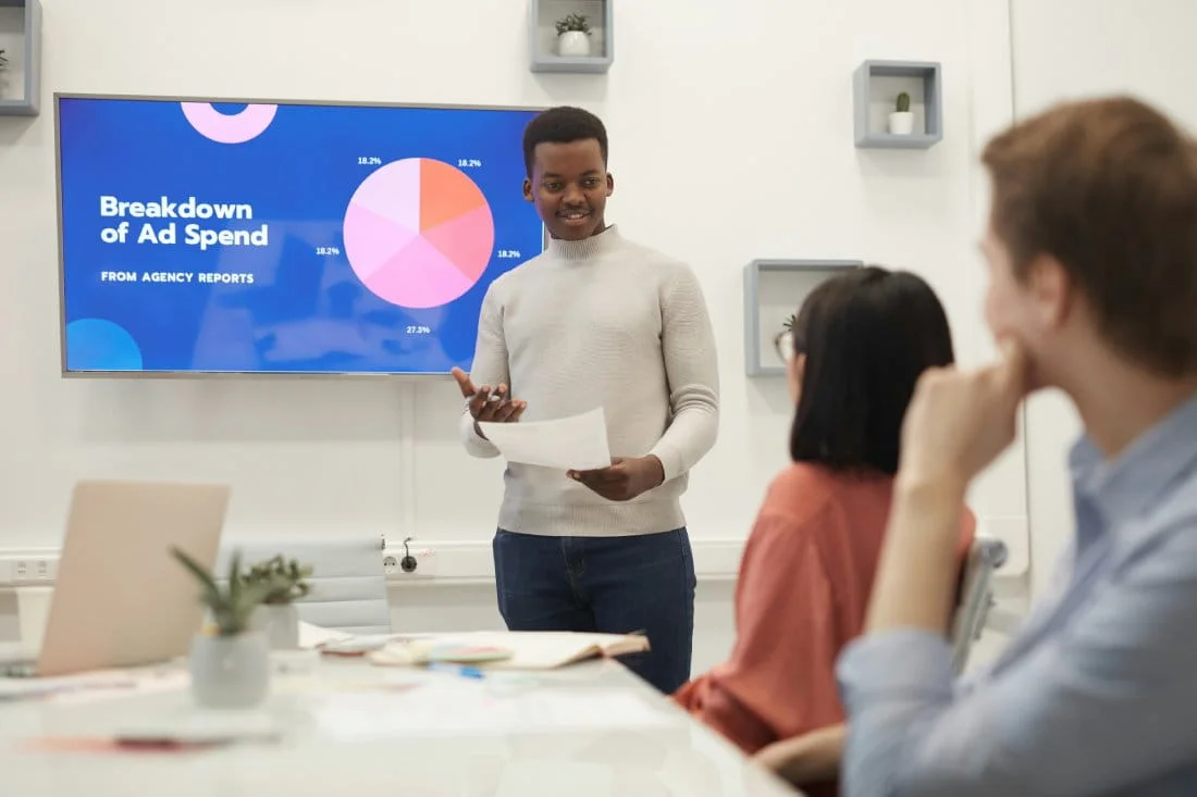 A man stands and presents data to two seated colleagues in a meeting room, using Simpl by Rob&Paul. Behind him, a large screen displays a slide with pie charts titled “Breakdown of Ad Spend.”.