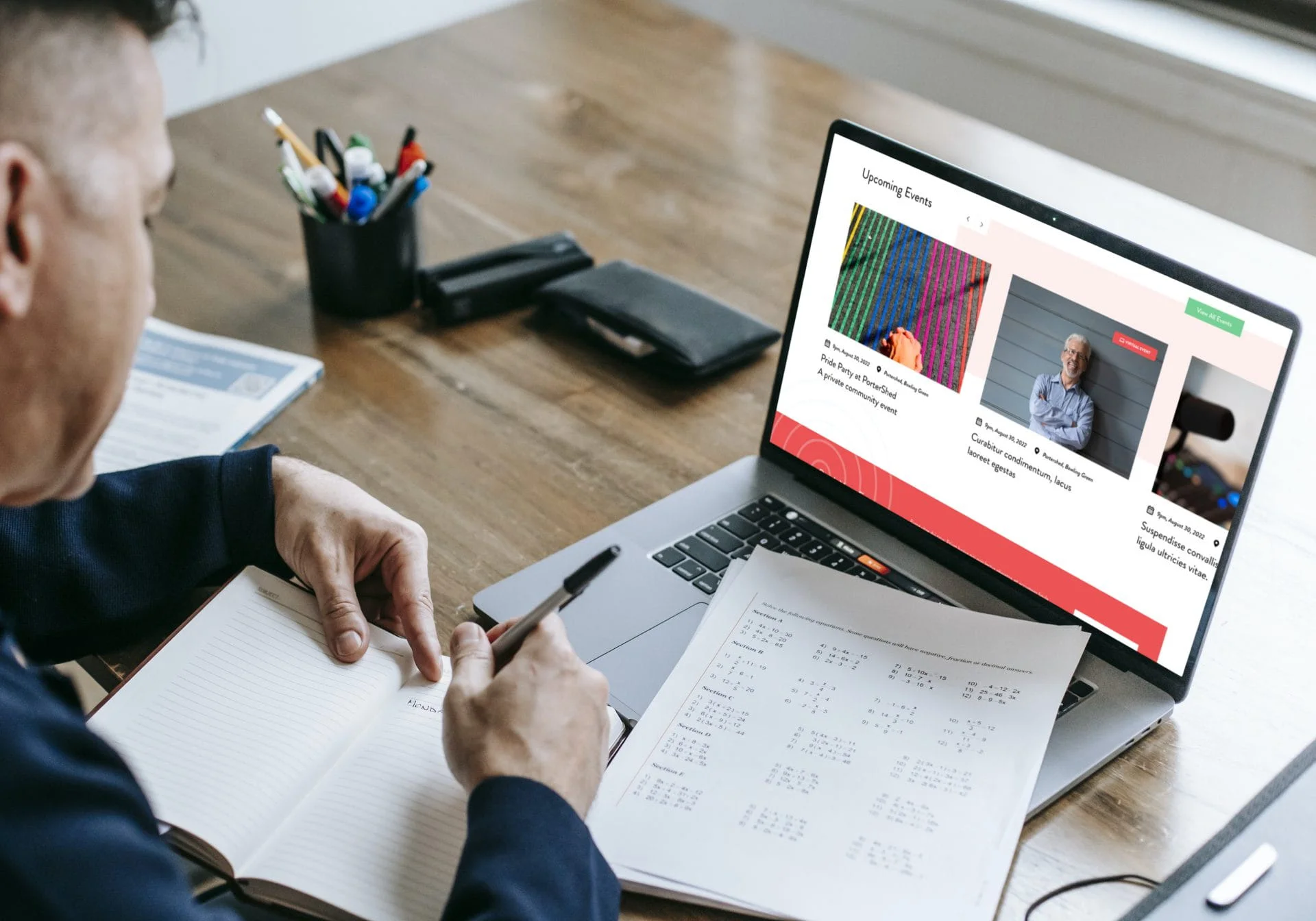 A person sits at a desk with a notebook and pen, looking at a laptop displaying upcoming events on Simpl by Rob&Paul. Papers with printed calendars and office supplies are also on the wooden desk.