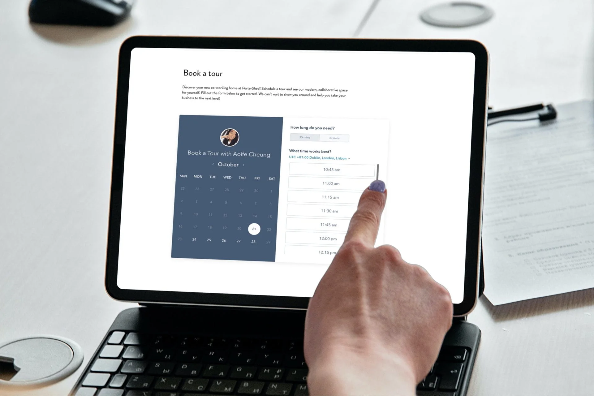 A person’s hand selects a time slot on a Simpl by Rob&Paul digital booking calendar displayed on a tablet screen, which is connected to a keyboard and placed on a desk.