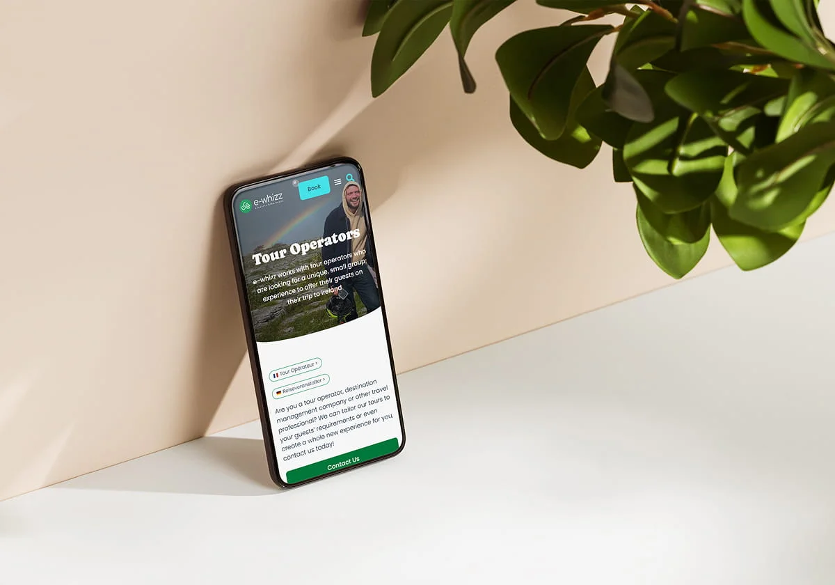 A smartphone leans against a beige wall on a white surface, displaying the Simpl by Rob&Paul Tour Operators webpage. Green leaves from a plant are visible in the upper right corner.