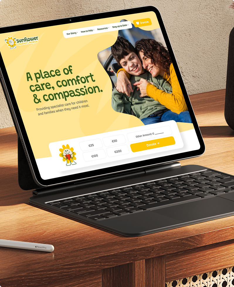 An open tablet on a desk displays the Simpl by Rob&Paul website with a yellow theme, showing a smiling woman hugging a laughing boy. Text reads: A place of care, comfort & compassion, and donation options are visible below.