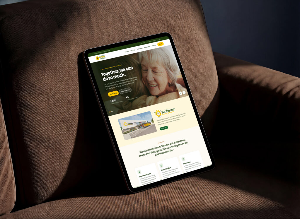 A tablet on a brown armchair displays a website with an elderly woman smiling and embracing someone, alongside information about a care facility called Sunflower Senior Living, highlighting their partnership with Mayo Roscommon Hospice Foundation.